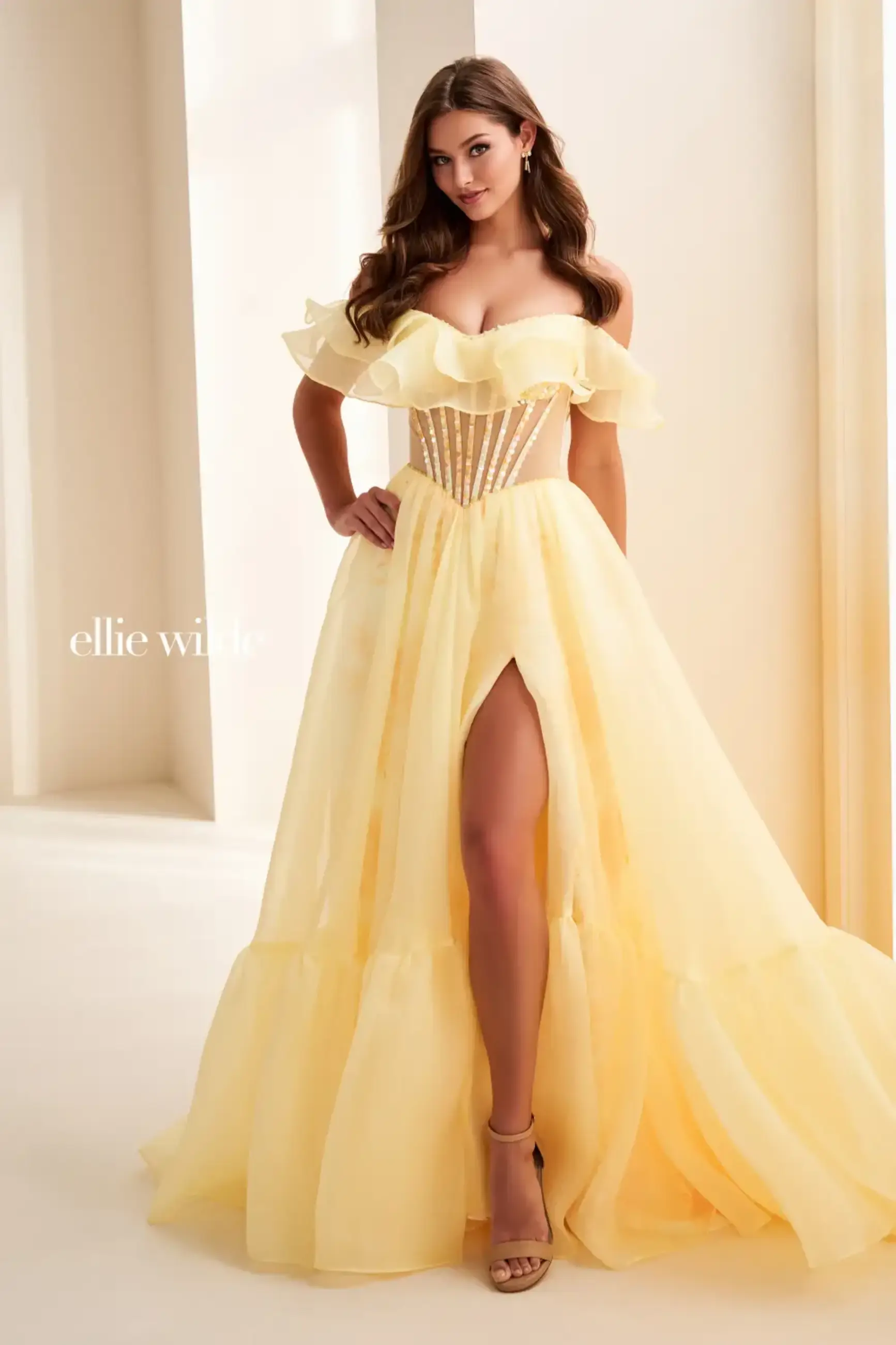 A woman wearing a strapless, off-the-shoulder yellow gown with ruffles and a high slit, showcasing a fitted bodice with a corset design. She has long, wavy hair and is posing elegantly.