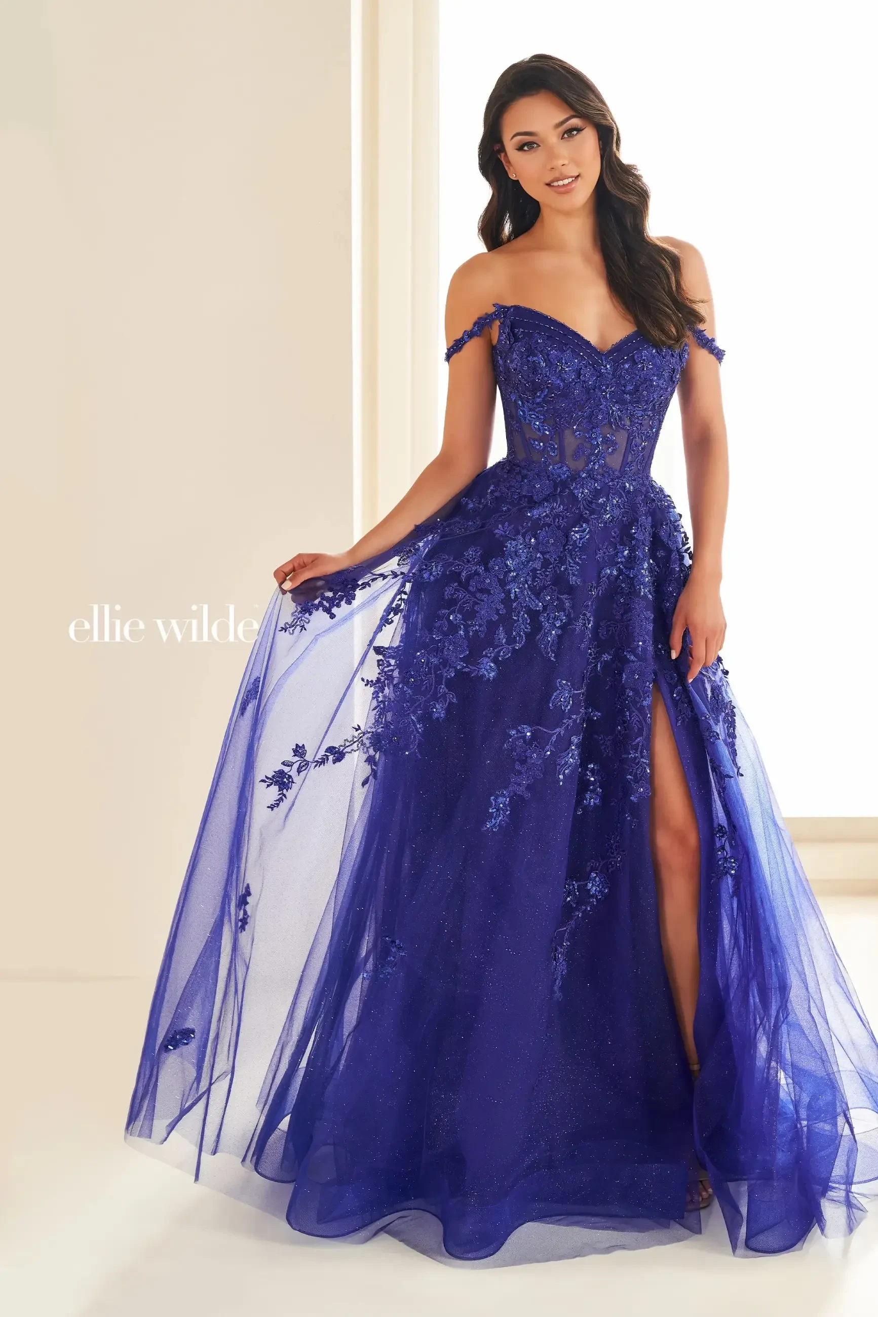 A woman in a vibrant, off-shoulder royal blue gown adorned with floral lace and sequins, featuring a high slit. She stands confidently, exuding elegance.