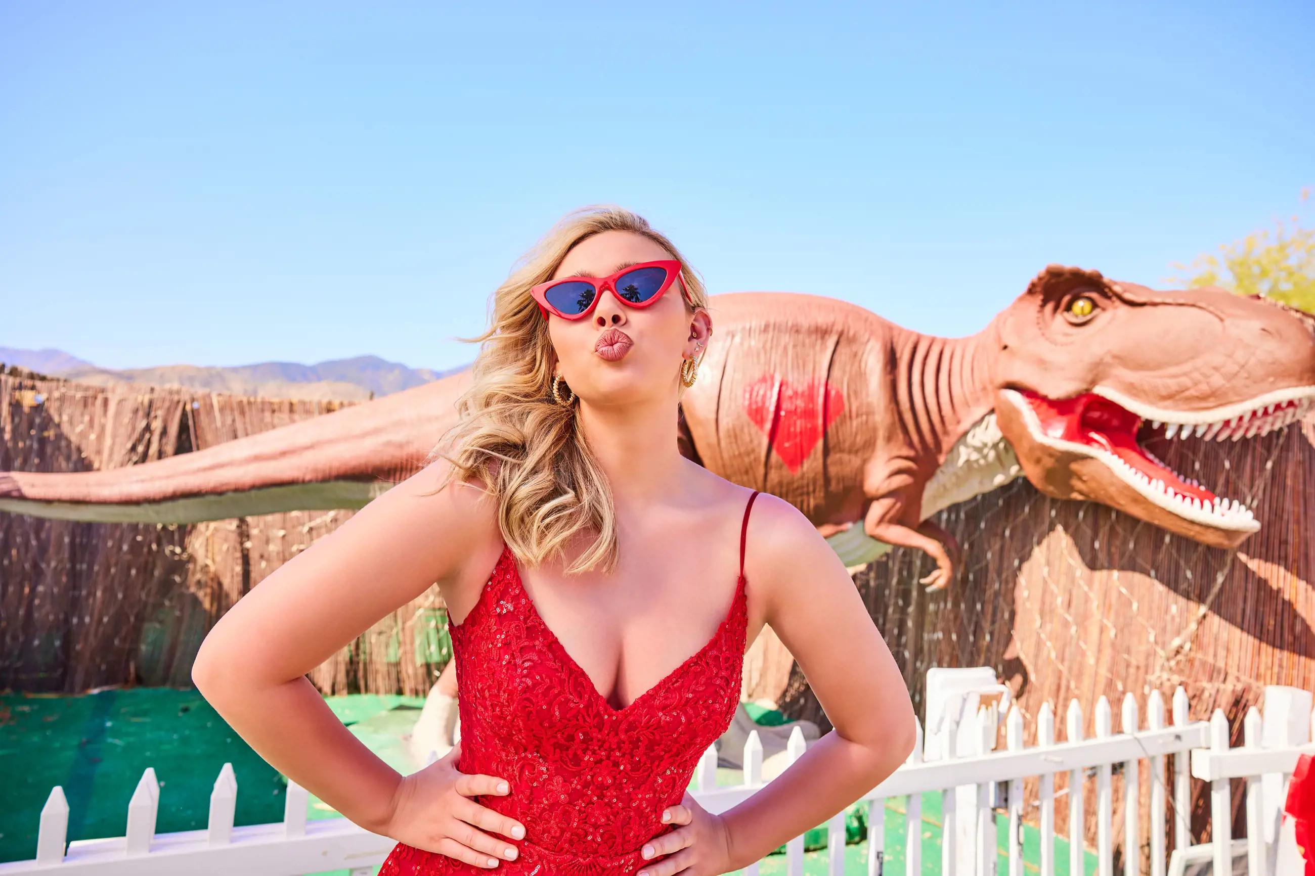 Blonde girl wearing red dress in front of huge t-rex plastic figure. Close-up